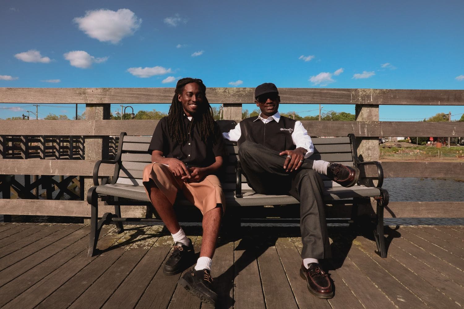 Two people seated on a bench on a wooden boardwalk on the Long Island waterfront, wearing Outskirtts